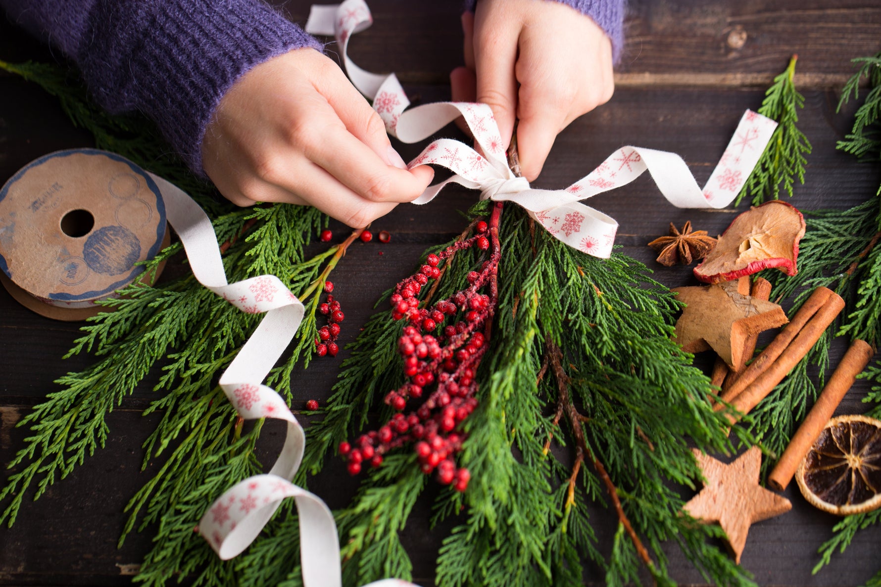 Woman's hands making a Holiday Swag out of cedar, berries and ribbon on a decorated craft table.